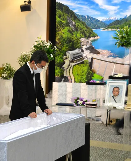 葬儀事例のサムネイル画像 黒部ダムの全景を写した祭壇の前でお父様に話しかけるご家族